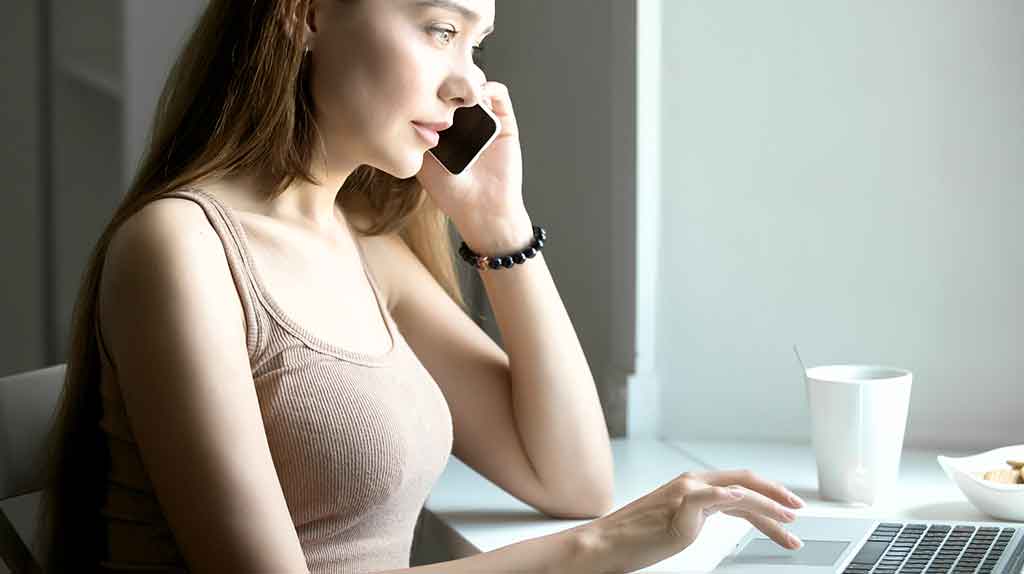Mulher num escritório com um smartphone na mão esquerda, um notebook a sua frente.