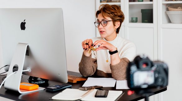 Mulher de óculos e cabelo curto, sentada em frente a uma mesa de escritório, segurando uma caneta nas mãos, olhando para um computador.