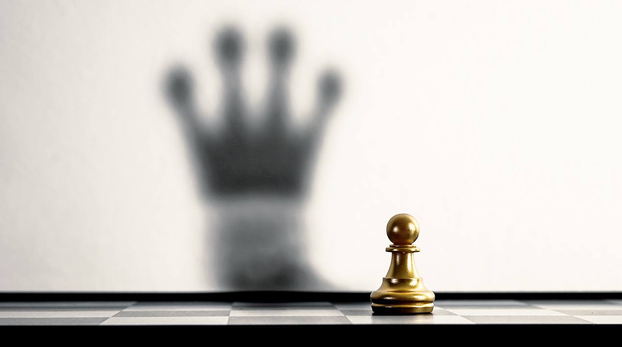 Pião do jogo de xadrez posicionado na casa preta com sombra projetada na parede no formato de coroa. 