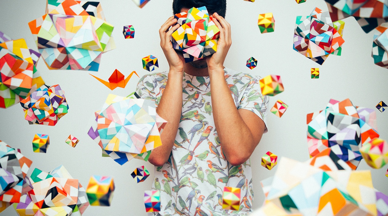 Homem segurando um origami a frente de seu rosto e circundado por diversos outros origamis. 