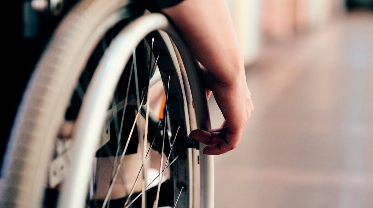 Close-up da roda lateral direita de uma cadeira de rodas e, sobre a roda, está apoiada a mão do cadeirante.