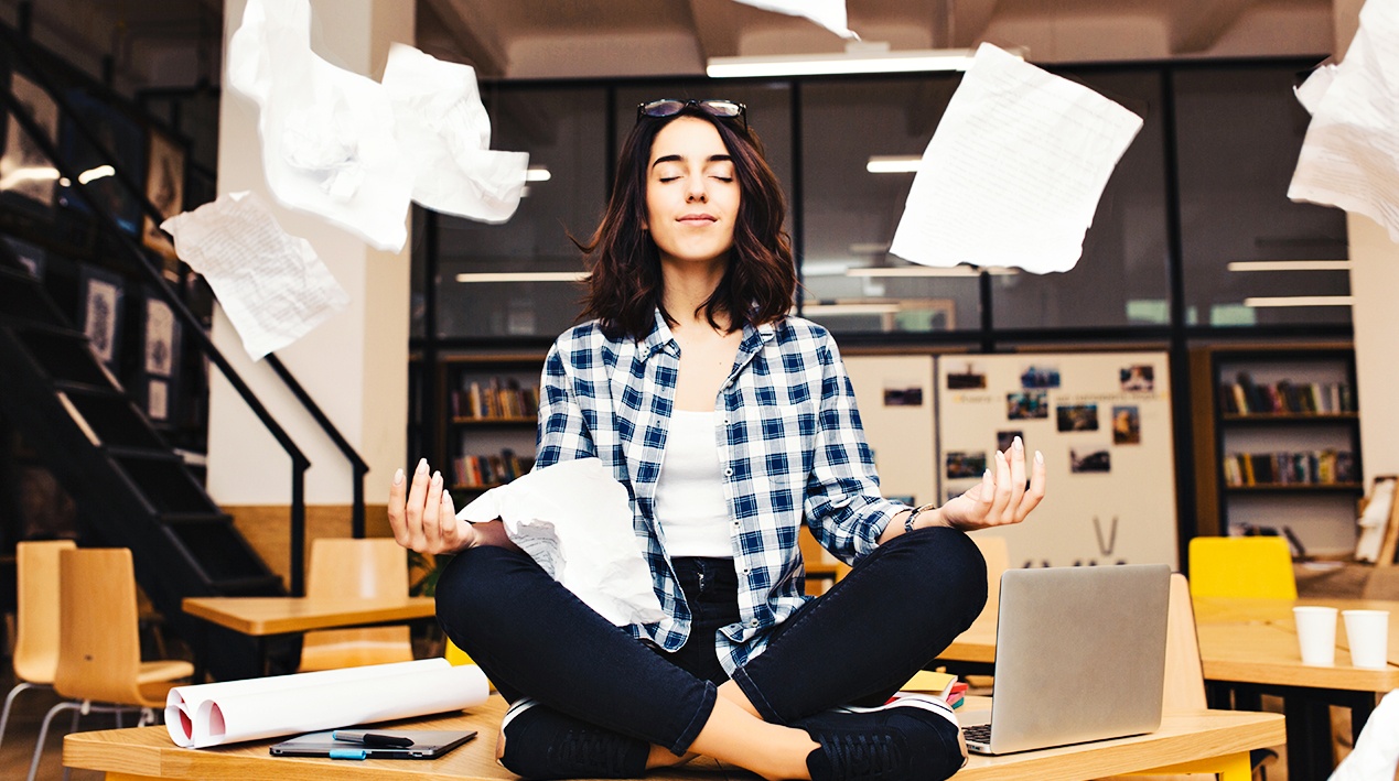 Mulher sentada em cima da mesa de um escritório, em posição de meditação, com papéis pairando no ar.