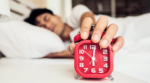 Homem deitado acordando ao som de um despertador, o qual ele tenta desligar.