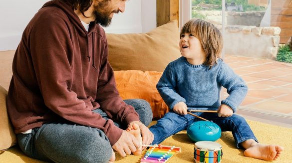 Homem adulto e uma criança sentados no tapete de uma sala. A criança está com baquetas nas mãos e em frente as pernas um tambor de brinquedo