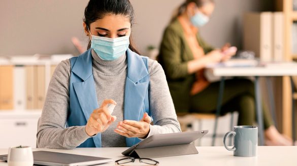 Mulher sentada em frente à uma mesa de escritório, higienizando as mãos com álcool spray, usando máscara de proteção compra a pandemia da COVID-19.