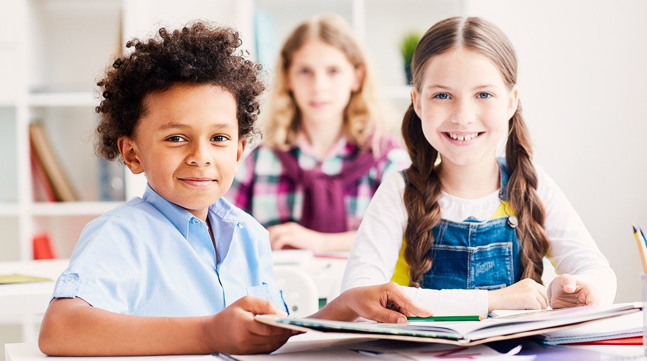 Três crianças em sala de aula, sendo um garoto e uma garota a frente, em que o garoto segura um caderno aberto e aponta para algo com o lápis. Todos estão sorrindo.