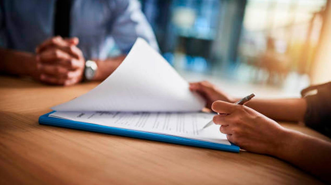 Close-up de duas pessoas com as mãos sobre uma mesa de escritório e uma delas está segurando uma caneta e avaliando um documento.