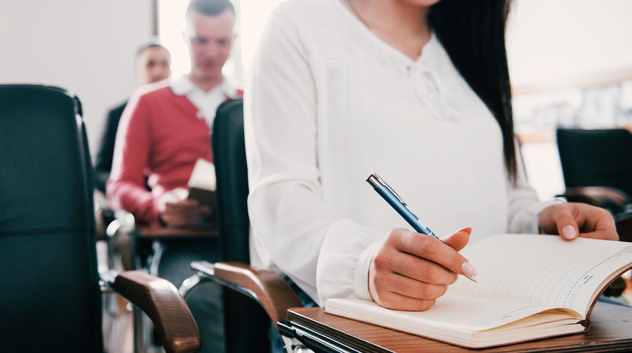 Estudantes do ensino médio em uma sala de aula. Detalhe em primeiro plano de uma aluna com uma agenda aberta e uma caneta na mão direita.