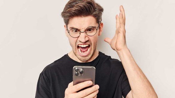 Homem com expressão de irritado, boa aberta, olhando para o smartphone que está em suas mãos.