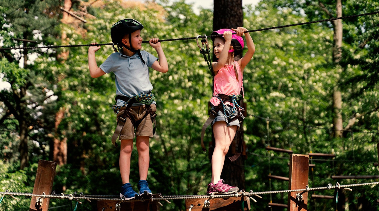 Crianças brincando numa floresta atravessando de um lado a outro por meio de um cabo de aço e aparatos de segurança.