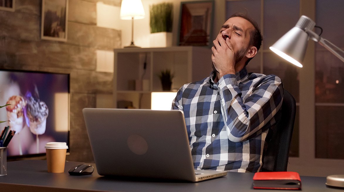 Homem em um escritório, sentado em a uma mesa de escritório frente e a um notebook, bocejando de sono.