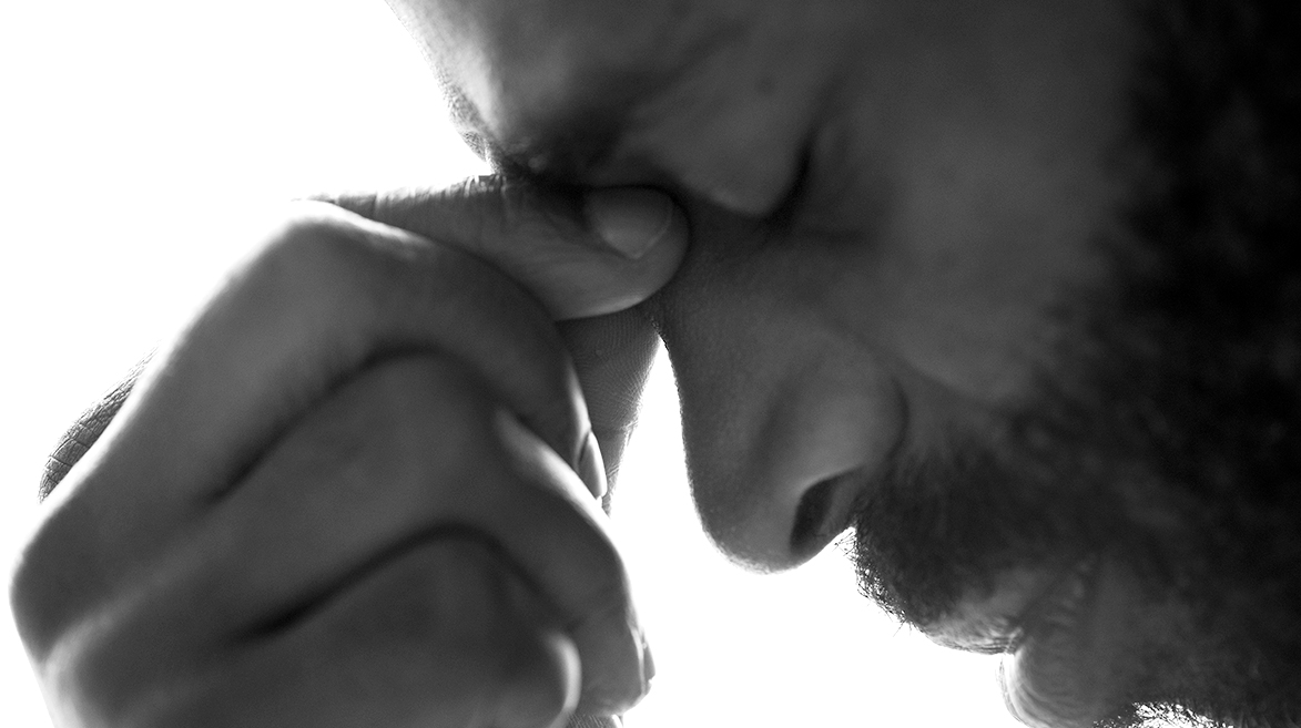 Homem de perfil com a cabeça levemente inclinada para baixo, com os dedos indicador e polegar segurando a parte do nariz que fica na altura dos olhos e com expressão de dor.